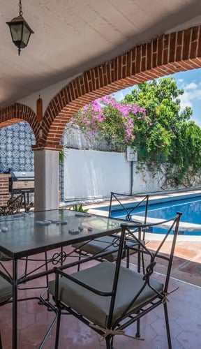 Covered patio with wrought-iron dining table framing a pool through a brick arch, with bougainvillea overhead